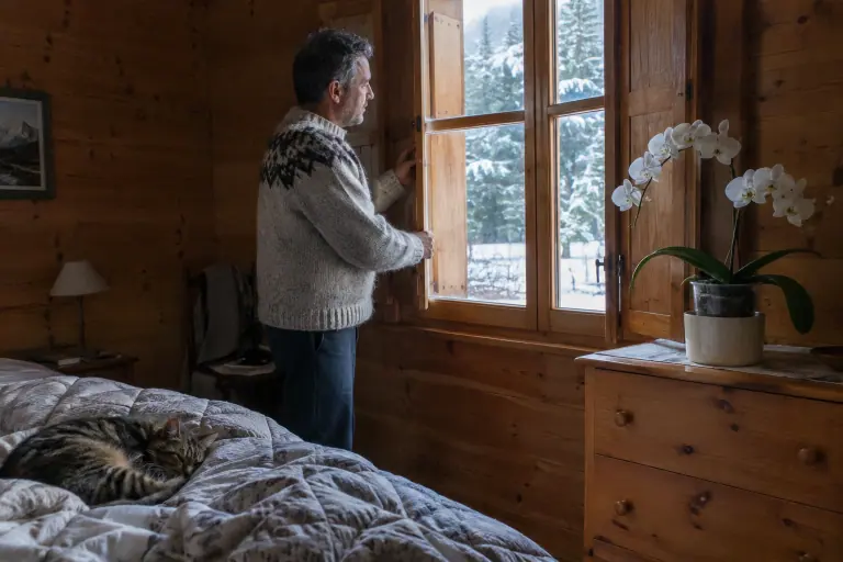 Dans une chambre de chalet savoyard, un homme ouvre les volets un matin de neige près d’une orchidée blanche posée sur une commode en pin
